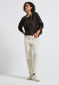 Veste bomber noire avec quincaillerie argentée, associée à un haut noir ajusté et un jean baggy beige clair. Des baskets blanches complètent la tenue.