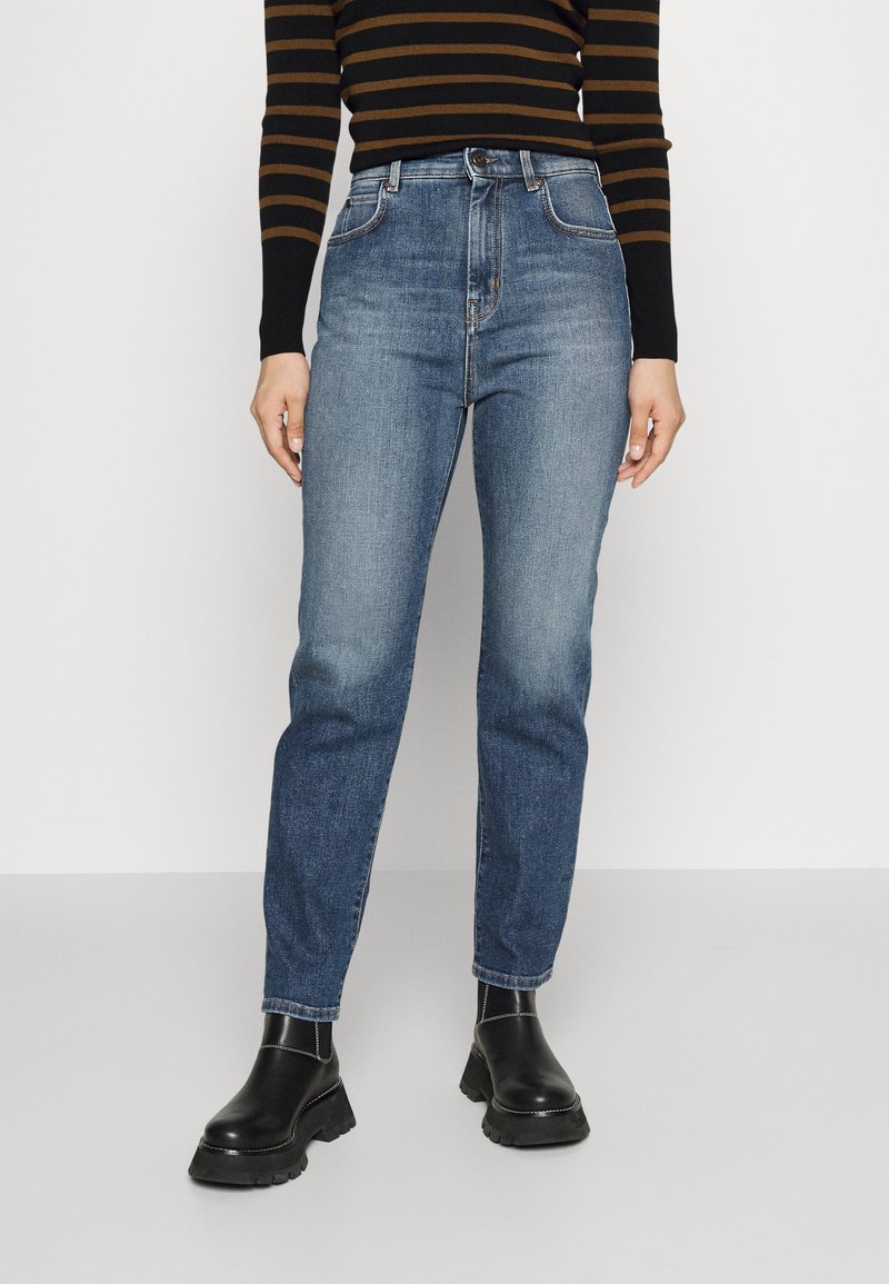 Femme portant un jean bleu taille haute avec un haut à manches longues à rayures foncées et des bottines noires épaisses sur un fond uni.
