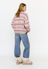 Pletený svetr s růžovými a béžovými vzory, volný střih, kombinovaný s světle modrými džínami a béžovými nadýchanými botami na platformě.