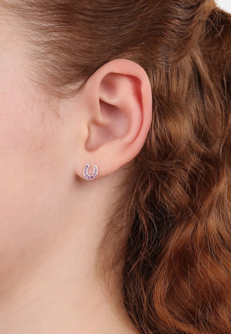 Pendientes en forma de herradura de color rosa con pequeñas piedras preciosas moradas, montadas en un metal de tono plateado, exhibidos en la oreja de un modelo contra un fondo de cabello rizado.