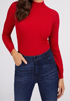 Femme portant un pull à col roulé rouge côtelé ajusté et un jean taille haute bleu foncé, une main dans la poche.