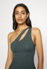 Asymmetrische one-shoulder jurk van donkergroene stof, nauwsluitend ontwerp met een gebogen halslijn en textuurdetails op de schouderband.