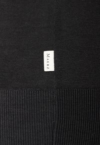 Svart stickad tröja med en slät textur och ribbad nederkant. Litet rektangulärt märke med "MAERZ" broderat på sidan.