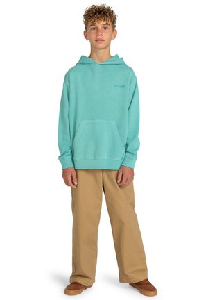 Sudadera turquesa con bolsillo frontal, puños acanalados y dobladillo; combinada con pantalones beiges de pierna ancha y zapatos marrones. Diseño de ajuste informal.
