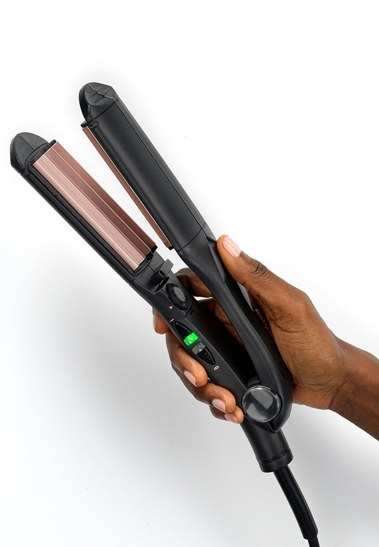 Plancha de pelo negra sostenida en la mano, con placas de oro rosa, pantalla digital, dial de control y un cable de alimentación largo.