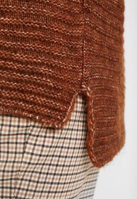 Brauner Strickpullover mit strukturiertem Finish, ausgestattet mit einem seitlichen Schlitz, kombiniert mit einem karierten Stoff in beigen und braunen Farbtönen.