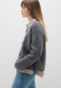 Cárdigan gris de punto con un diseño texturizado, con cierres de botones. Lleva puesto sobre una camisa a cuadros, combinado con jeans azules.