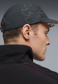 Sort cap med mønstret skygge, der har et subtilt grafisk design på toppen. Modellen har en sort jakke med røde detaljer på.