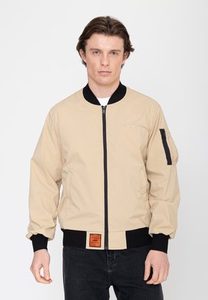 Jonge man draagt beige bomberjack met zwarte kraag, manchetten en rits, poserend tegen een witte achtergrond.