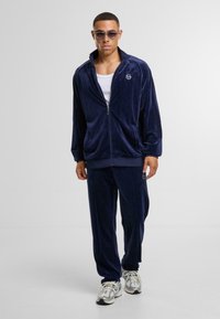 Chándal de felpa de terciopelo azul marino con cremallera y pantalones a juego, que presenta un logo blanco, puños acanalados y tela texturizada. Se lleva con gafas y zapatillas.