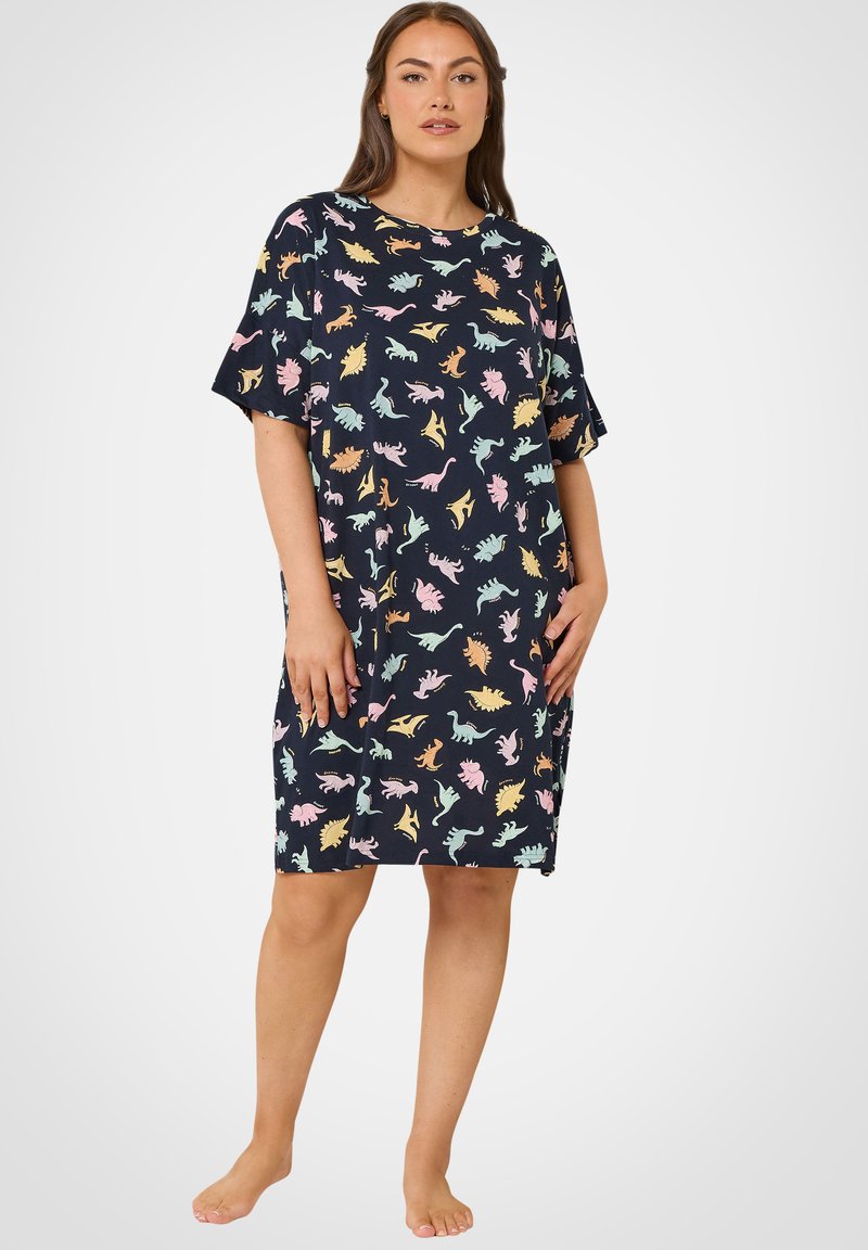 Frau trägt ein kniegesäumtes marineblaues Kleid mit kurzen Ärmeln und bunten Dinosauriermustern in Pastellfarben, steht barfuß vor einem weißen Hintergrund.