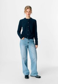 Cardigan marine avec un col rond et cinq boutons, associé à un jean large bleu clair et des chaussures noires. Tissu doux, coupe décontractée.