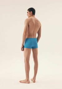 Niebieskie kąpielówki wykonane z elastycznego materiału, z dopasowanym krojem i gładką fakturą. Model pokazany z tyłu, na bosaka na neutralnym tle.