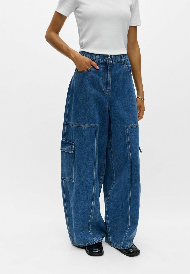 Personne portant un pantalon cargo en denim bleu à jambes larges avec de grandes poches latérales, un haut blanc à manches courtes et des chaussures noires, debout devant un fond uni.