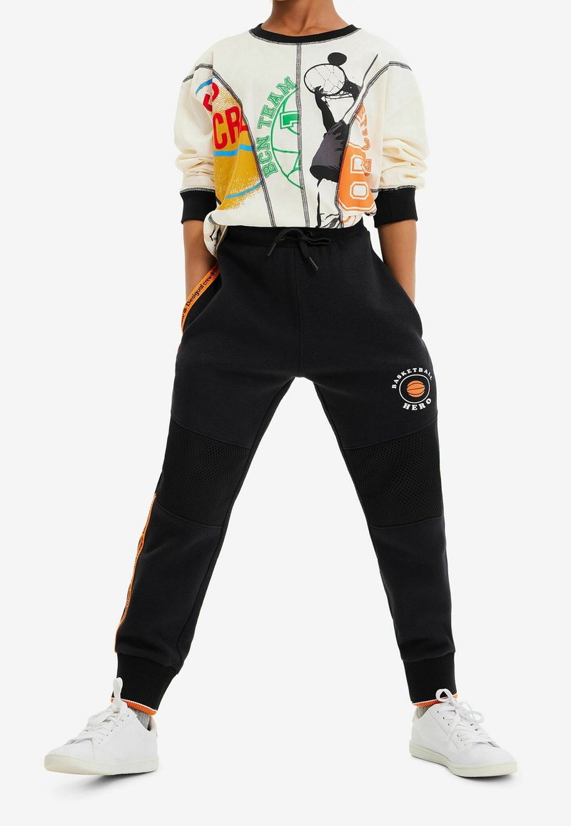Witte sweatshirt met grafische print, zwarte joggers met gaaspanelen en oranje accenten, gecombineerd met witte sneakers.