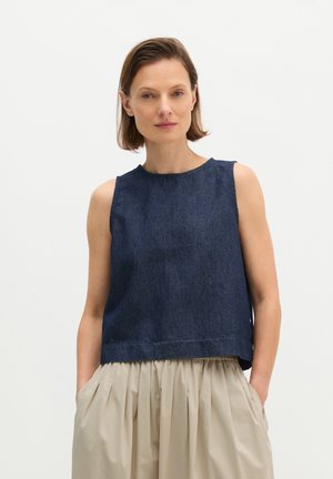 Femme aux cheveux bruns courts porte un haut en jean bleu foncé sans manches et une jupe plissée beige, debout les mains dans les poches de la jupe.