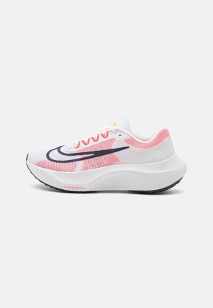 Chaussure de course blanche et rose avec une semelle épaisse tachetée et un logo Nike swoosh noir et rouge sur le côté sur un fond uni.