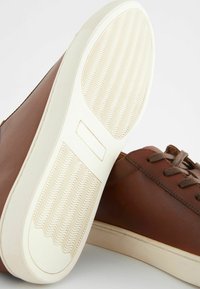 Bruine leren sneakers met een gladde afwerking, voorzien van bruine veters en een gestructureerde beige rubberen zool met een golvend patroon.