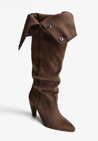 Brązowy zamszowy kozak do kolan z odwróconym mankietem, srebrnymi zatrzaskami, spiczastym czубkiem i smukłym wysokim obcasem. Miękka tekstura i wyrafinowany design.