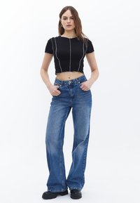 Schwarzes Cropped-Top mit kontrastierendem weißen Nähten und lockeren blauen Jeans mit mittlerem Bund und ausgestellten Beinen. Das Model steht mit den Händen in den Hüften.
