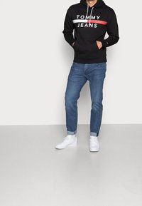 Černá mikina s kapucí s logem "TOMMY JEANS" v bílé a červené barvě, klokanková kapsa, spárovaná s ohrnutými modrými džíny a bílými teniskami.