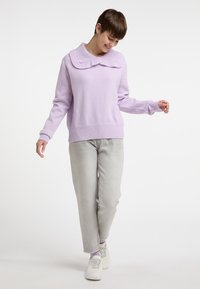 Maglione lavorato a maglia color lavanda chiaro con un ampio colletto, polsini e orlo a coste. Indossato con pantaloni grigi chiari e scarpe da ginnastica bianche.