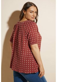Blusa rossa a fantasia con scollatura arricciata e maniche corte. Presenta pieghe verticali e un design morbido. Indossata con jeans blu.