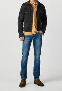 Black denim jacket, mustard polo, blue jeans, brown boots. Jacket features front pockets, button closure, and a relaxed fit.