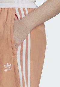 Meruňkové sportovní šortky s bílým třemi pruhy, elastickým pasem a boční kapsou. Obsahují vyšívané logo Adidas. Hladký povrch.
