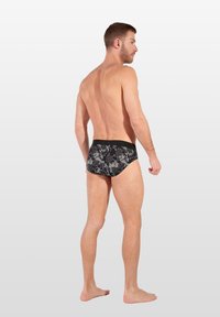 Schwarze Schwimmslips mit floralem Muster, glatter Textur und kontrastierendem Bund, seitlich mit neutralem Hintergrund gezeigt.