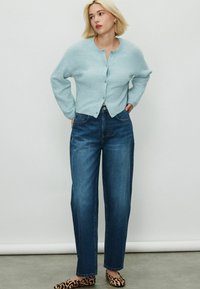 Lichtblauwe gebreide cardigan, ronde hals, knoopsluiting aan de voorkant, gecombineerd met hoog-waist blauwe jeans en loafers met luipaardprint. Soepele textuur.
