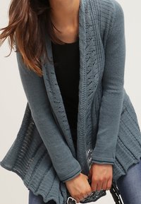 Öppen cardigan i texturerad blå stickning med detaljerade mönster och ribbad f åll. Har långa ärmar och en flödande design.