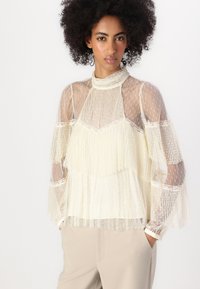 Sheer ivory blouse with textured patterns, pleated details, and flared sleeves. High neck design with polka dot accents and lace trim.