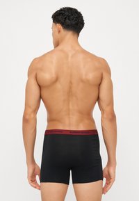 Schwarze Boxer Shorts mit burgunderfarbenem Bund, der mit dem Logo "BJÖRN BORG" versehen ist, aus glattem Stoff und in einer taillierten Passform. Sicht von hinten.