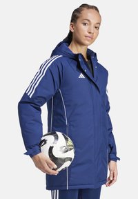 Marineblaue, wasserdichte Jacke mit weißen Akzenten, Reißverschluss, Kapuze und verstellbaren Bündchen. Eine Person hält einen Schwarz-Weiß-Fußball.