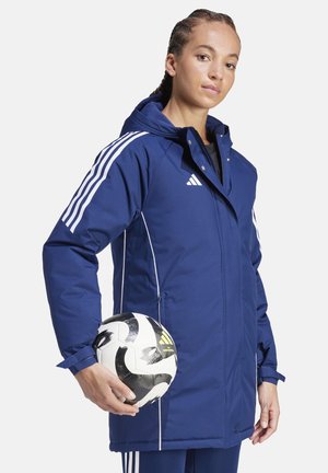 Marineblaue, wasserdichte Jacke mit weißen Akzenten, Reißverschluss, Kapuze und verstellbaren Bündchen. Eine Person hält einen Schwarz-Weiß-Fußball.