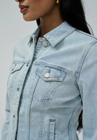 Veste en denim bleu clair avec col, poches avant et boutons en métal argenté. Affiche une texture décolorée et un design classique.