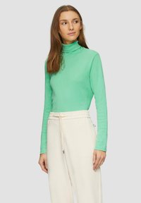 Langärmliges Turtleneck-Oberteil aus lebhaftem mintgrünem Stoff, kombiniert mit cremefarbenen, Jogginghosen mit Kordelzug. Glatte Textur, figurbetontes Design.