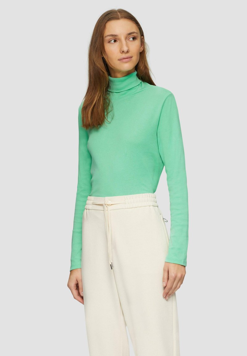 Langärmliges Turtleneck-Oberteil aus lebhaftem mintgrünem Stoff, kombiniert mit cremefarbenen, Jogginghosen mit Kordelzug. Glatte Textur, figurbetontes Design.