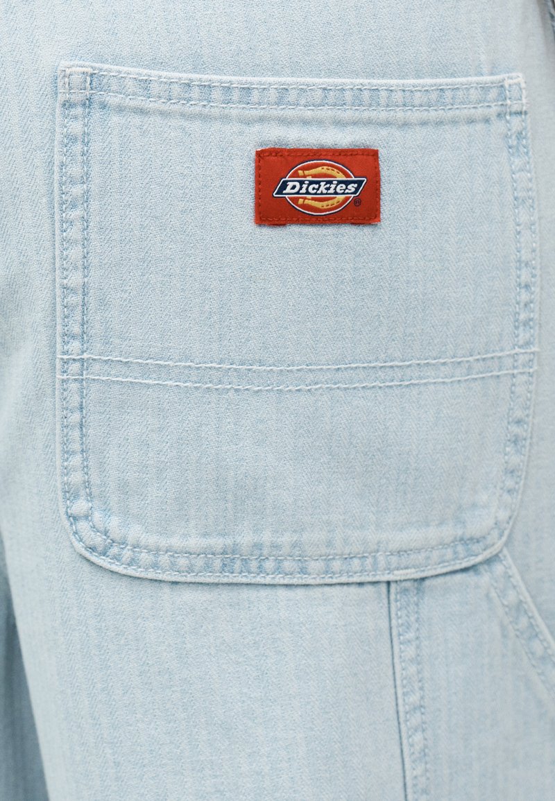 Tessuto di denim azzurro chiaro con una grande tasca cucita che presenta un'etichetta rettangolare rossa con il logo Dickies in bianco e giallo.