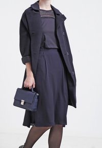 Námořnický kabát s texturovaným povrchem, přetíraný přes ladící blůzu a sukni, doplněný malou, strukturovanou kabelkou.
