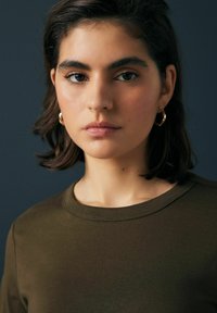 Grünes kurzärmliges Hemd mit rundem Ausschnitt, getragen mit goldenen Creolen. Glatte Textur, minimalistisches Design, Nahaufnahme der Gesichtszüge.