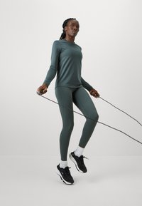 Športový outfit pozostávajúci z tmavozelenej dlhoprstej blúzky, zhodných legín a čiernych tenisiek s bielymi akcentmi. V rukách držiaca skákačku.