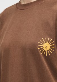 Καφέ πουκάμισο με μικρό κίτρινο γραφικό ήλιου που εμφανίζει ένα ήρεμο πρόσωπο και ακτίνες που ακτινοβολούν στην αριστερή πλευρά του στήθους.