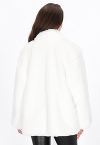 Vrouw met lang bruin haar, gekleed in een witte donzige jas en zwarte leren broek, gezien van achteren tegen een witte achtergrond.