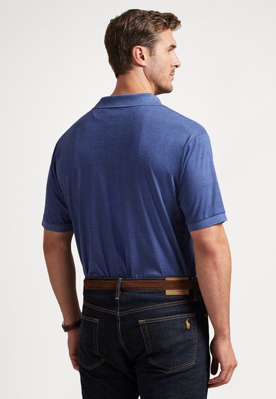 Άνδρας που φορά μπλε πουκάμισο πόλο με καρό σχέδιο και κοντά μανίκια, μπαίνει μέσα σε σκούρο τζιν παντελόνι με καφέ ζώνη, κοιτάζοντας προς τα πίσω πάνω σε απλό φόντο.