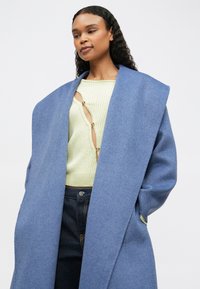 Nő hosszú kék gyapjú kabátot visel, alatt világoszöld kötött felső, kivágásokkal és gombokkal, sötétkék farmernadrággal, a kezét a zsebében tartja.