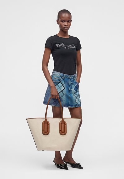 Γυναίκα με μαύρο t-shirt Marc Jacobs και τζιν φούστα κρατά μία μεγάλη μπεζ τσάντα ώμου με καφέ λαβές μπροστά σε μονόχρωμο φόντο.