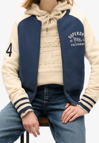 Granatowa i kremowa kurtka typu bomber zapinana na zamek, z prążkowanymi mankietami, ozdobiona nadrukiem graficznym i kontrastowym kolorem rękawów. Noszona na beżowej bluzie z kapturem.