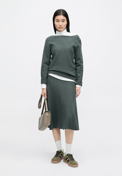 Γκρίζα πλεκτή μπλούζα και ταιριαστή φούστα μέχρι το γόνατο, τοποθετημένη πάνω από λευκό ριχτό πουλόβερ. Ολοκληρωμένο με ελαιόχρωμα αθλητικά παπούτσια και γκρι τσάντα.